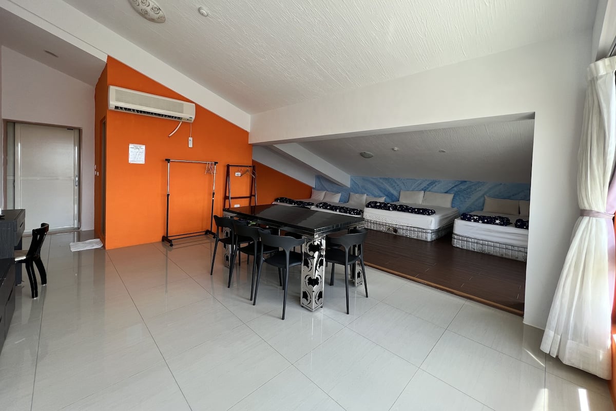 Una habitación amplia con una mesa larga y sillas, una cama con cabecera azul y una pared de acento naranja.