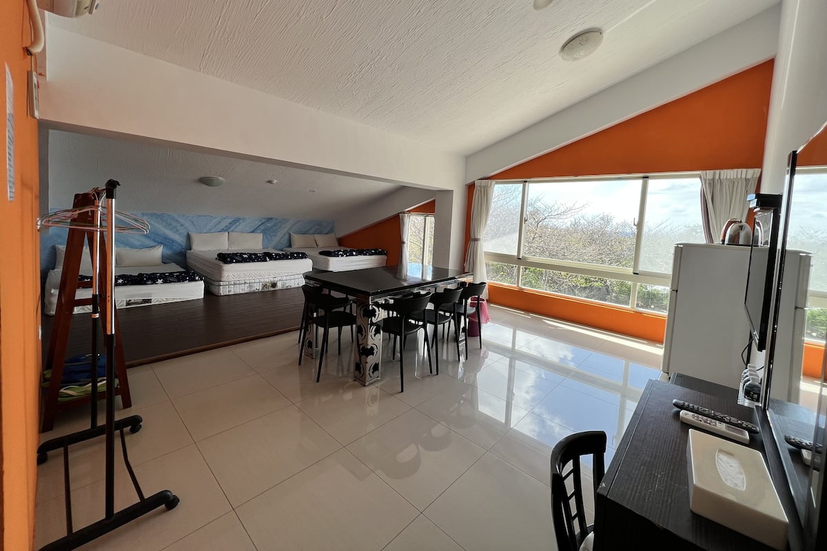 Amplia habitación con techo alto, zona de comedor con sillas y mesa, un conjunto de sofás y un ventanal con vistas a árboles.