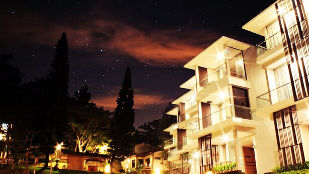 Un edificio moderno de varios pisos con balcones y ventanas amplias, rodeado de un jardín bien iluminado.