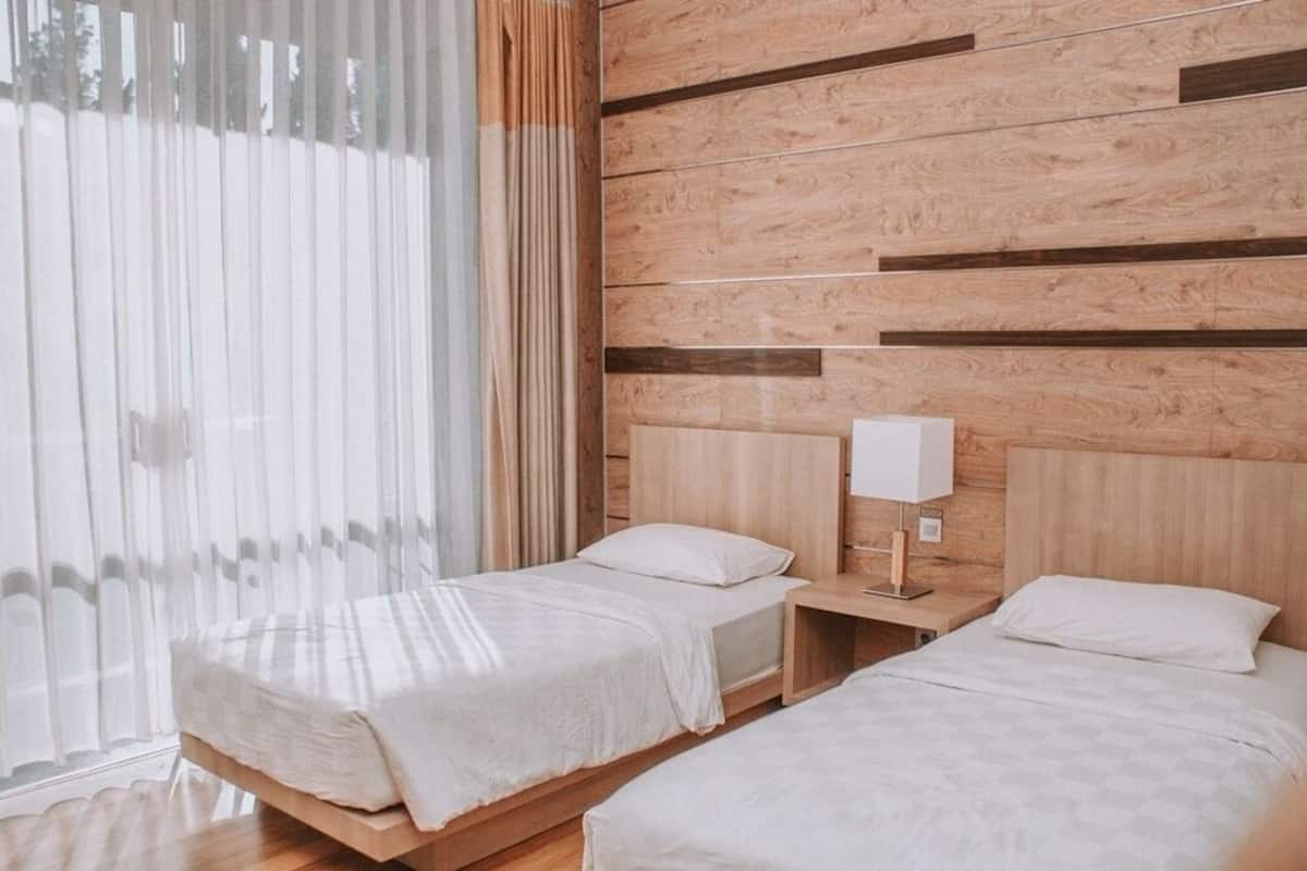 Ein Hotelzimmer mit zwei Betten, Holz-Kopfteilen, einem Nachttisch mit Lampe und durchsichtigen Vorhängen.