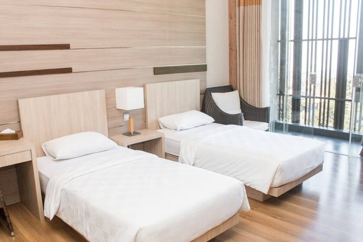 Ein Hotelzimmer mit zwei Betten, einem Nachttisch mit Lampe, einem Korbstuhl und einem großen Fenster mit Blick.