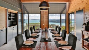 A dining area with a long table set for a meal, large windows offering a view of the sea, and a wooden ceiling. - Te Whau Lodge (Waiheke Island)