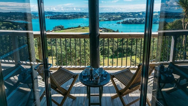 A balcony with a table set for two, overlooking a scenic view of a lake and surrounding landscape. - Te Whau Lodge (Waiheke Island)