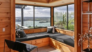 A cozy cabin with a large window offering a scenic view of a lake and hills. - Te Whau Lodge (Waiheke Island)