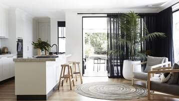A modern kitchen with white cabinetry, wooden stools, and a large island. There is a dining area with a round rug and a sofa.