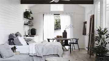 A cozy, well-lit room with a bed, a small table, and chairs. There are plants on shelves and a window with curtains.