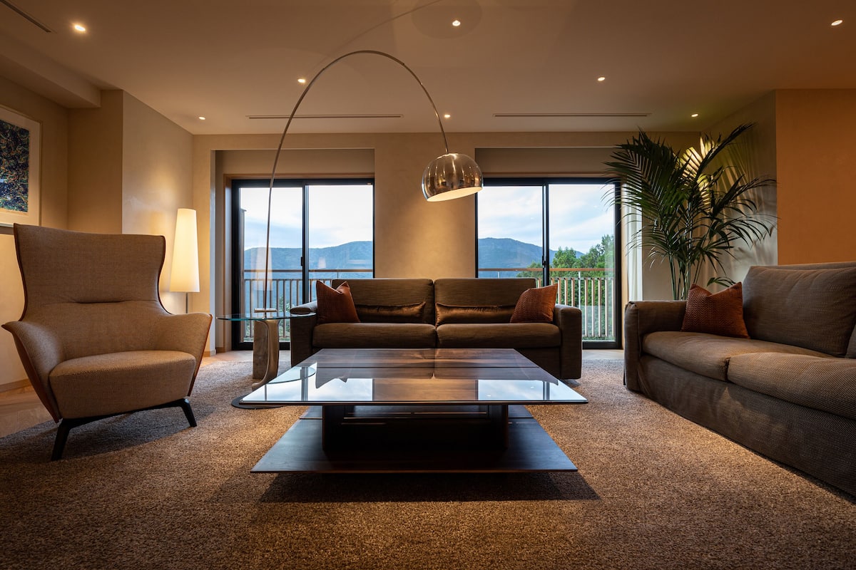 Ein modernes Wohnzimmer mit einem Glastisch, zwei Sofas, einem Sessel und einer großen Pflanze.
