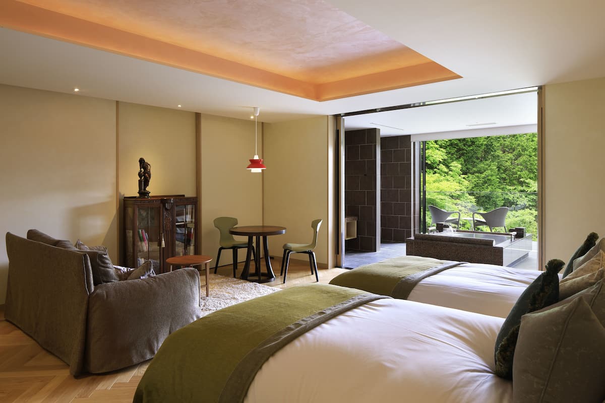Ein modernes Hotelzimmer mit zwei Betten, einer Couch, einem Esstisch und Stühlen. Ein großes Fenster bietet Blick auf Grünflächen außerhalb.