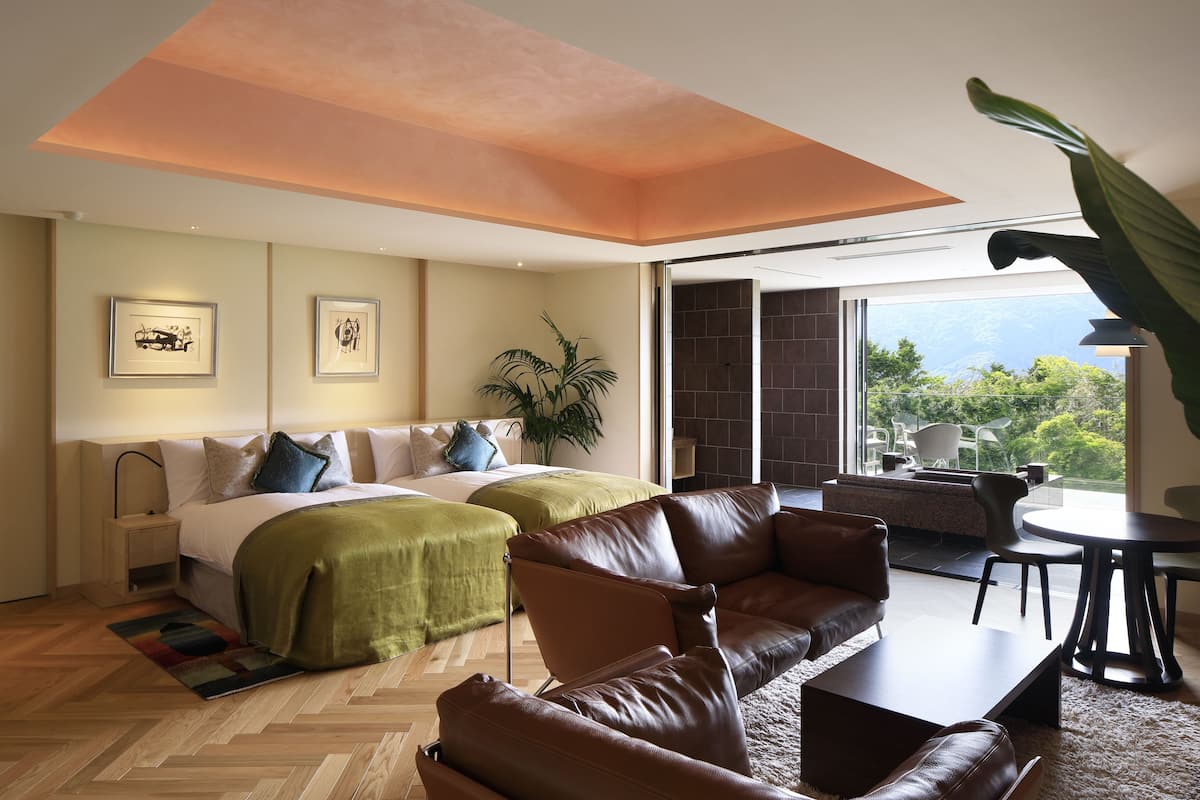 Ein modernes Hotelzimmer mit Bett, einer Couch, einem Esstisch und einem großen Fenster mit Blick ins Freie.
