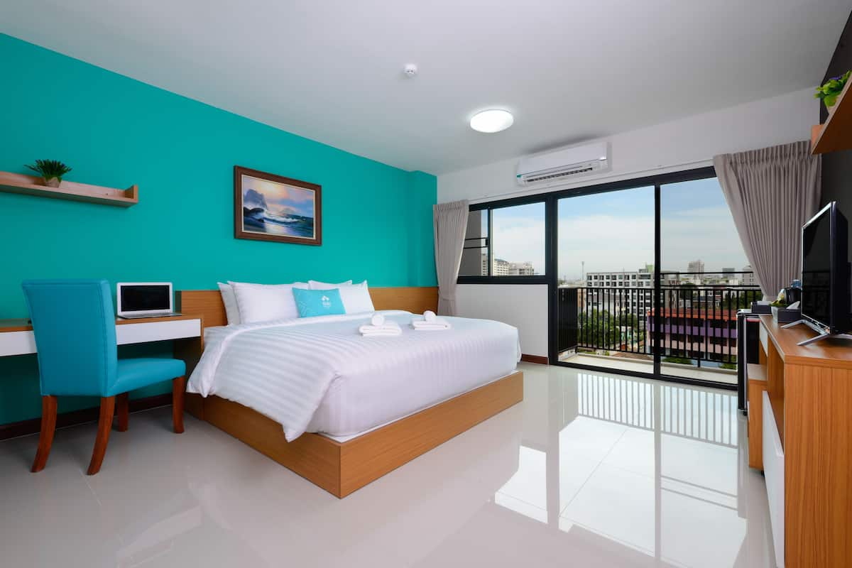 A modern hotel room with a large bed, a desk, a laptop, and a view of the city through a large window.