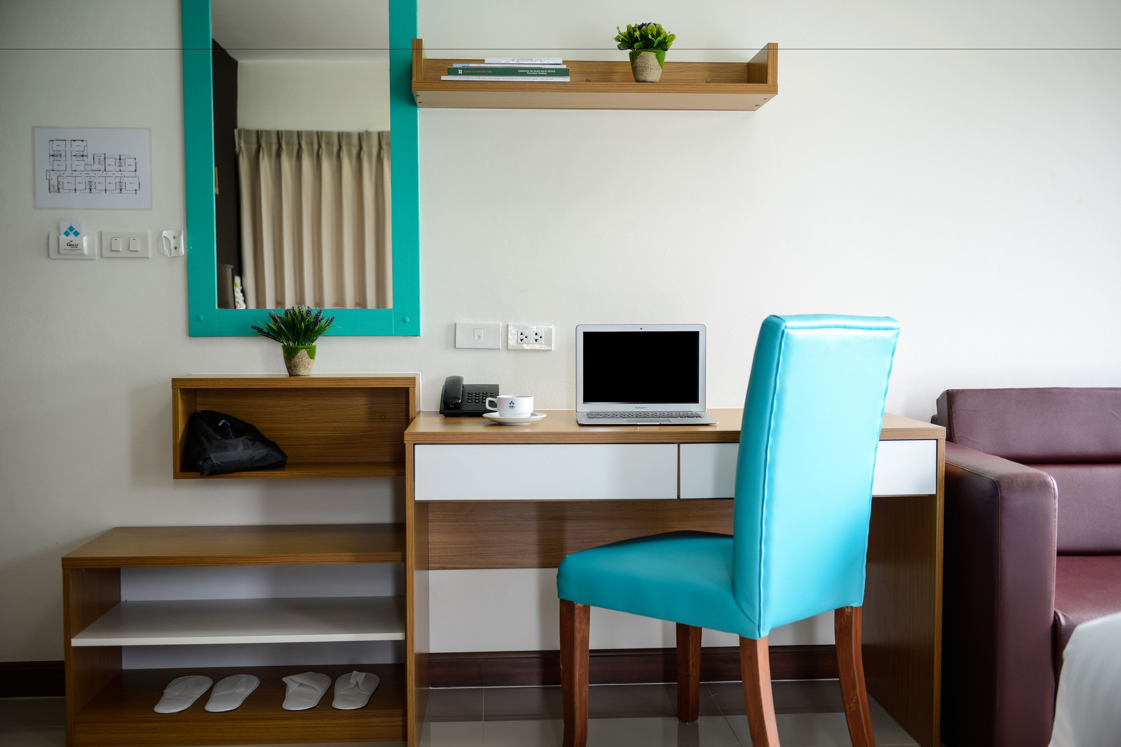 Une chambre d’hôtel avec un bureau en bois, un ordinateur portable, une chaise de couleur turquoise, une petite étagère avec une plante, une paire de chaussons et un miroir.