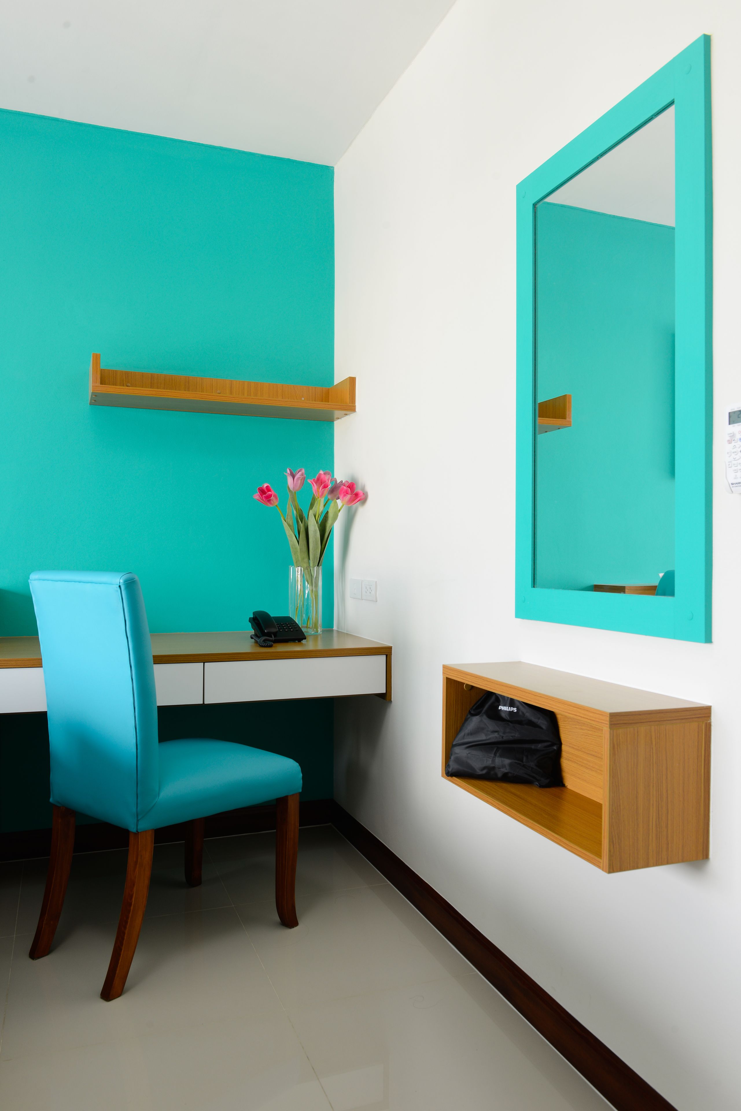 A teal-colored room with a wooden desk, a blue chair, a mirror, and a vase with pink flowers.