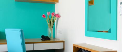 A teal-colored room with a wooden desk, a blue chair, a mirror, and a vase with pink flowers.