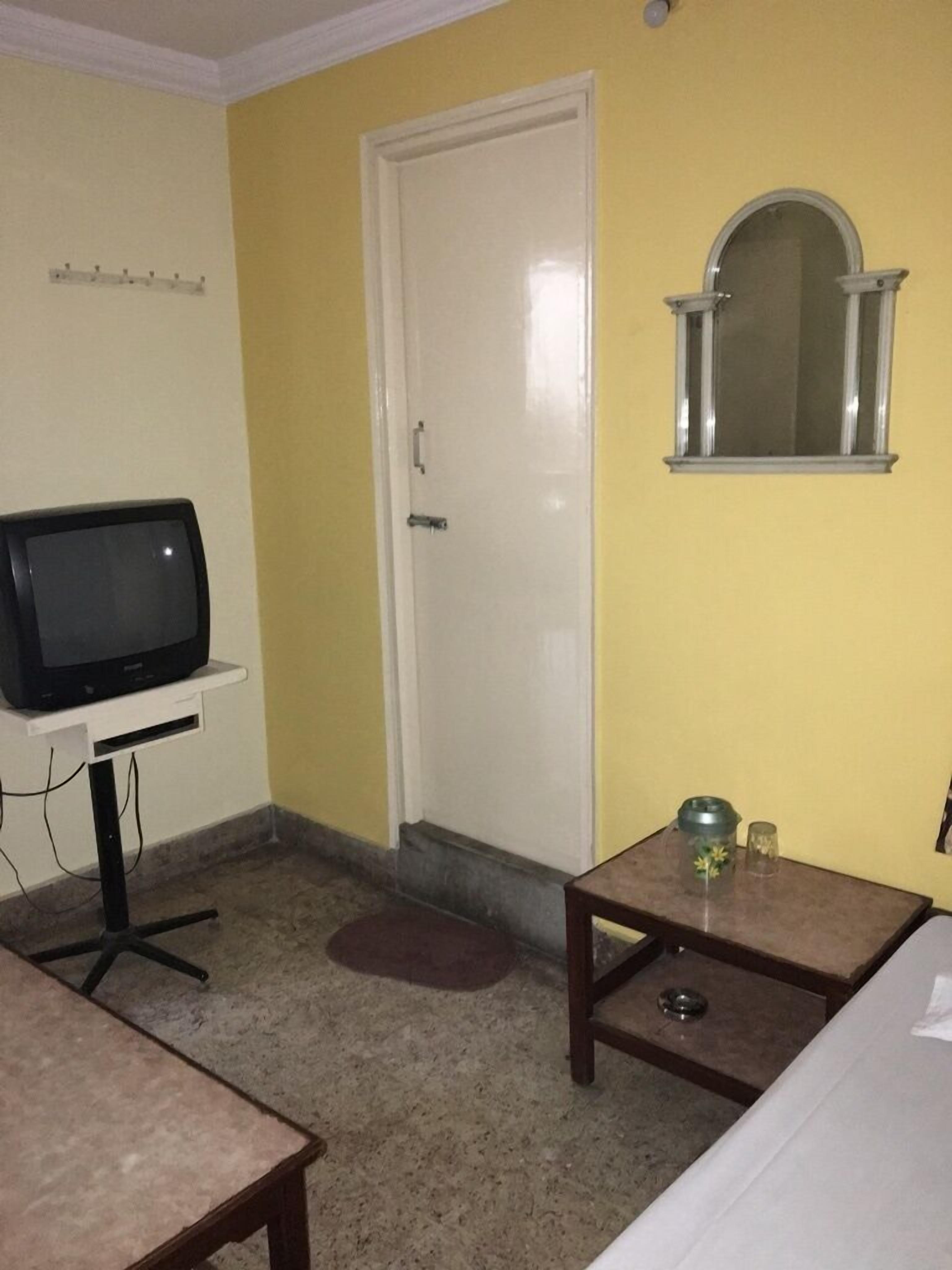 A small room with a television on a stand, a door, a small table with a glass and a plant, and a bed.