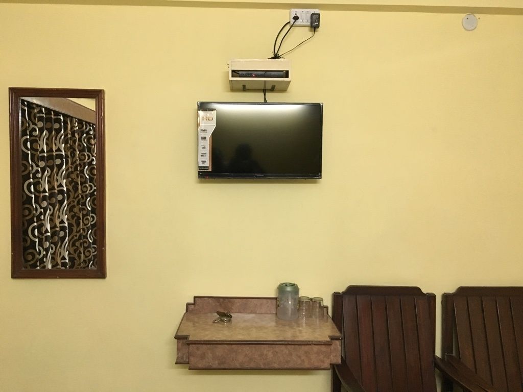 A room with a mounted flat-screen TV, a decorative wall piece, a small shelf with a container, and a wooden chair.