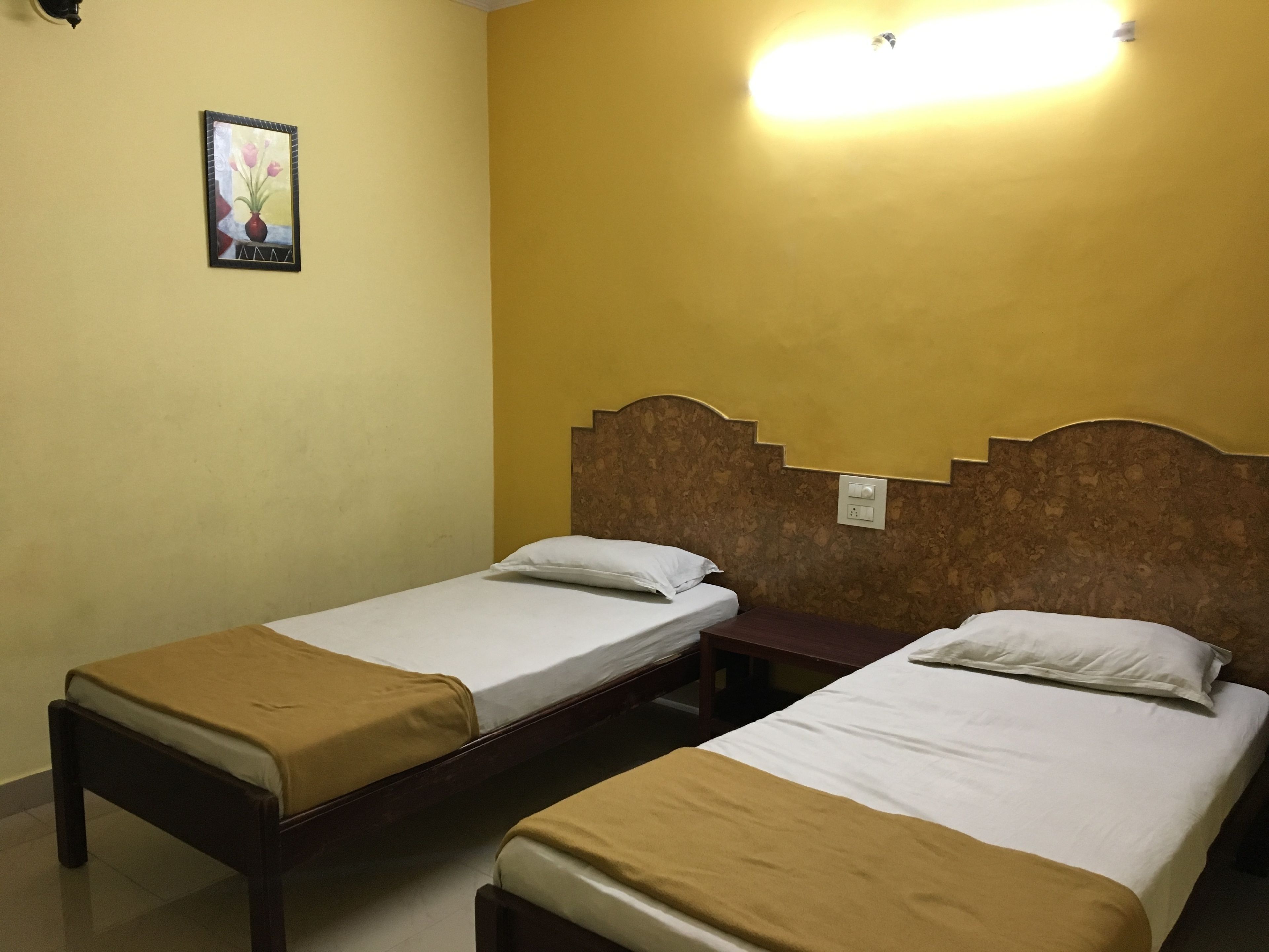 A room with two single beds, each with a brown headboard and white bedding. There is a small table between the beds, and a wall-mounted light fixture above it.