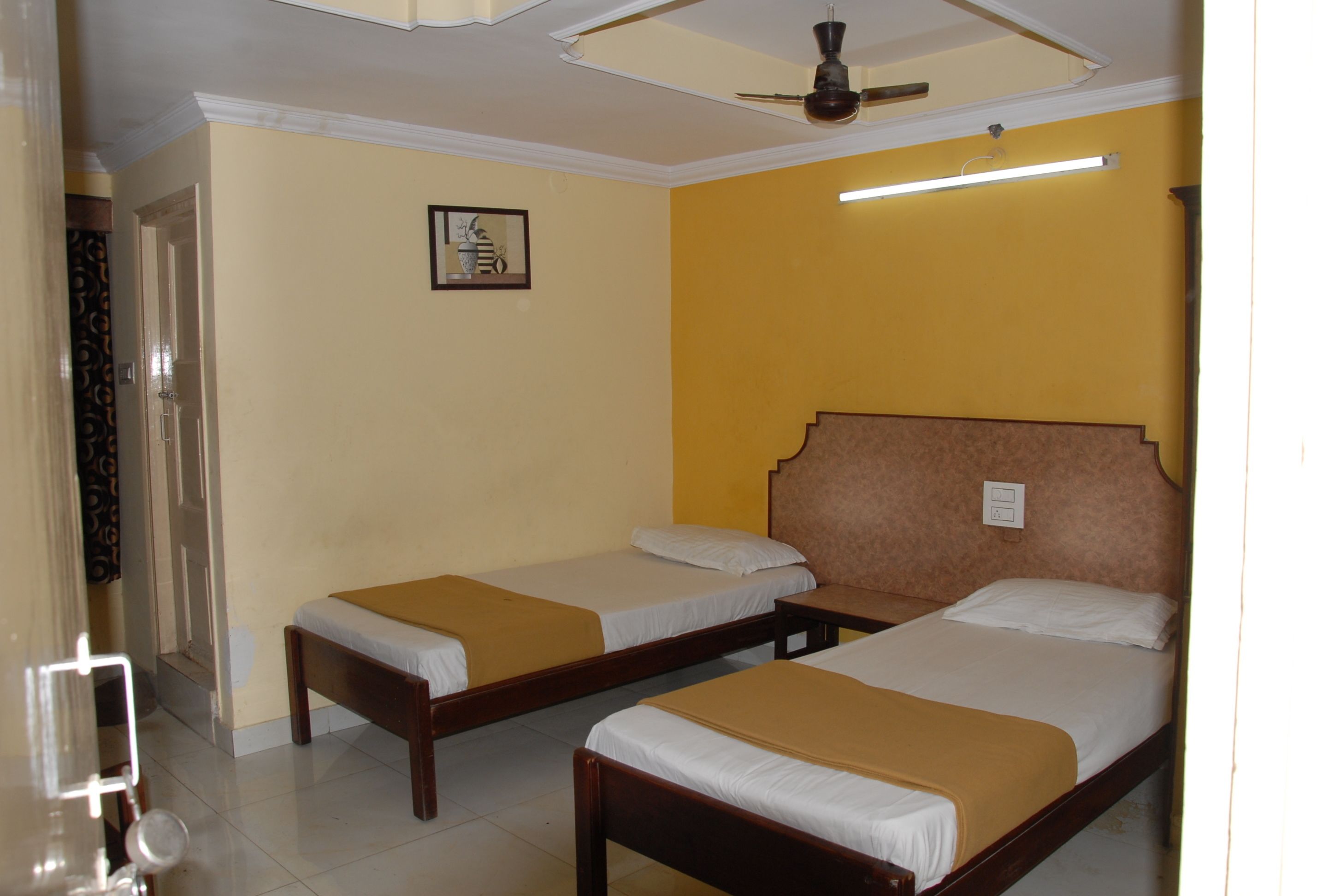 A hotel room with two single beds, a ceiling fan, a framed picture on the wall, and a light fixture above the beds.