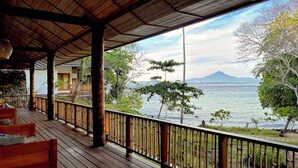 Una terraza de madera con barandilla de bambú que da a un cuerpo de agua y una montaña al fondo.