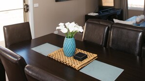A dining area with a dark wooden table, four chairs, a vase with white flowers, and a television on a black stand. - Bell's Cottage Collective (West Bendigo)