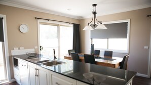 A modern kitchen with a large island, double sink, and a dining area with a table and chairs. - Bell's Cottage Collective (West Bendigo)