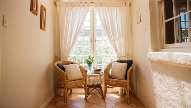 A corner room with two wicker chairs, a small table with books and a vase, and a window with white curtains. - Bell's Cottage Collective (West Bendigo)