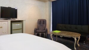 A hotel room with a bed, a television on a cabinet, a chair, a small table with a green folder, and a large window with curtains. - Hotel Shalala (Okinawa)