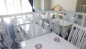 A dining area with white ornate chairs and tables, a blue carpet, and a view of the outside through large windows. - Hotel Shalala (Okinawa)
