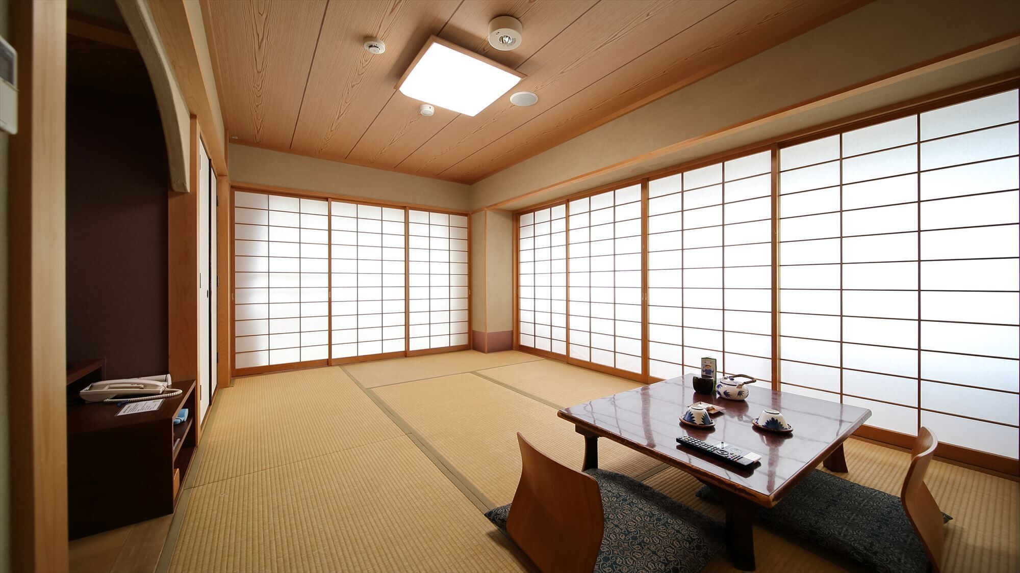 A traditional Japanese room with tatami flooring, sliding shoji doors, a low table with a remote control, and a small cabinet with a telephone.