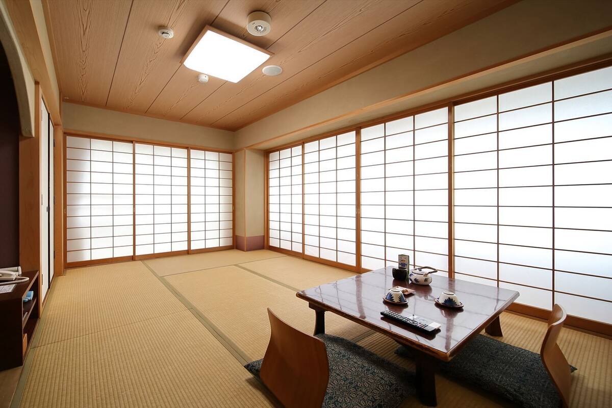 Una habitación japonesa tradicional con suelo de tatami, puertas correderas shoji, una mesa baja con un control remoto y un armario pequeño con un teléfono.