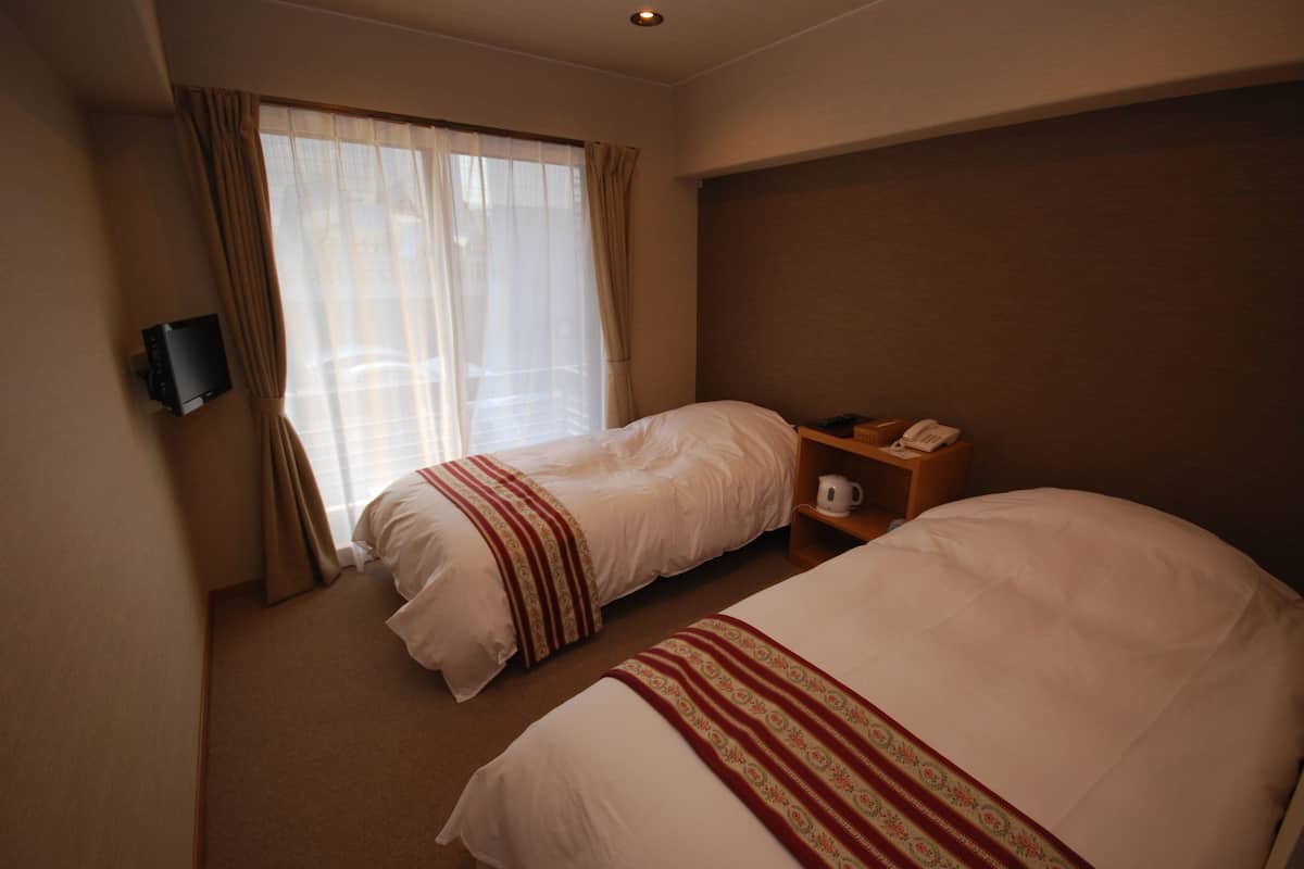 A hotel room with two beds, a TV mounted on the wall, a bedside table with a phone and a kettle, and a window with curtains.