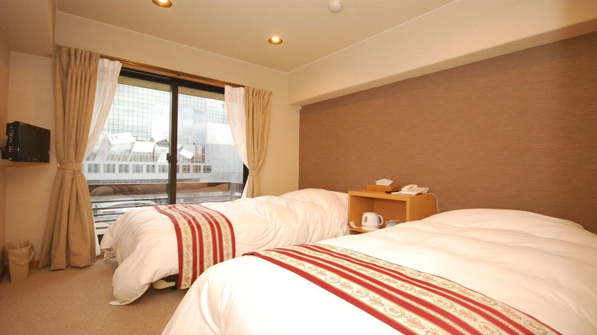 A hotel room with two beds, a television, a bedside table with a phone and a kettle, and a view of a modern building through the window.