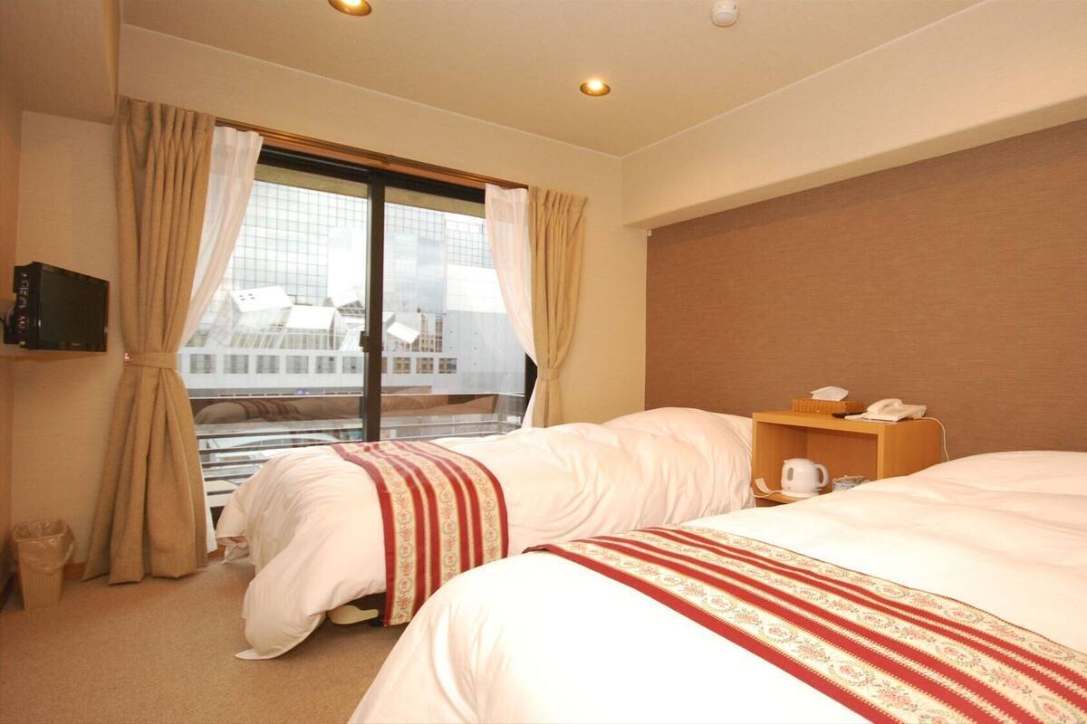 Una habitación de hotel con dos camas, un televisor, una mesita de noche con teléfono y hervidor, y vista a un edificio moderno a través de la ventana.
