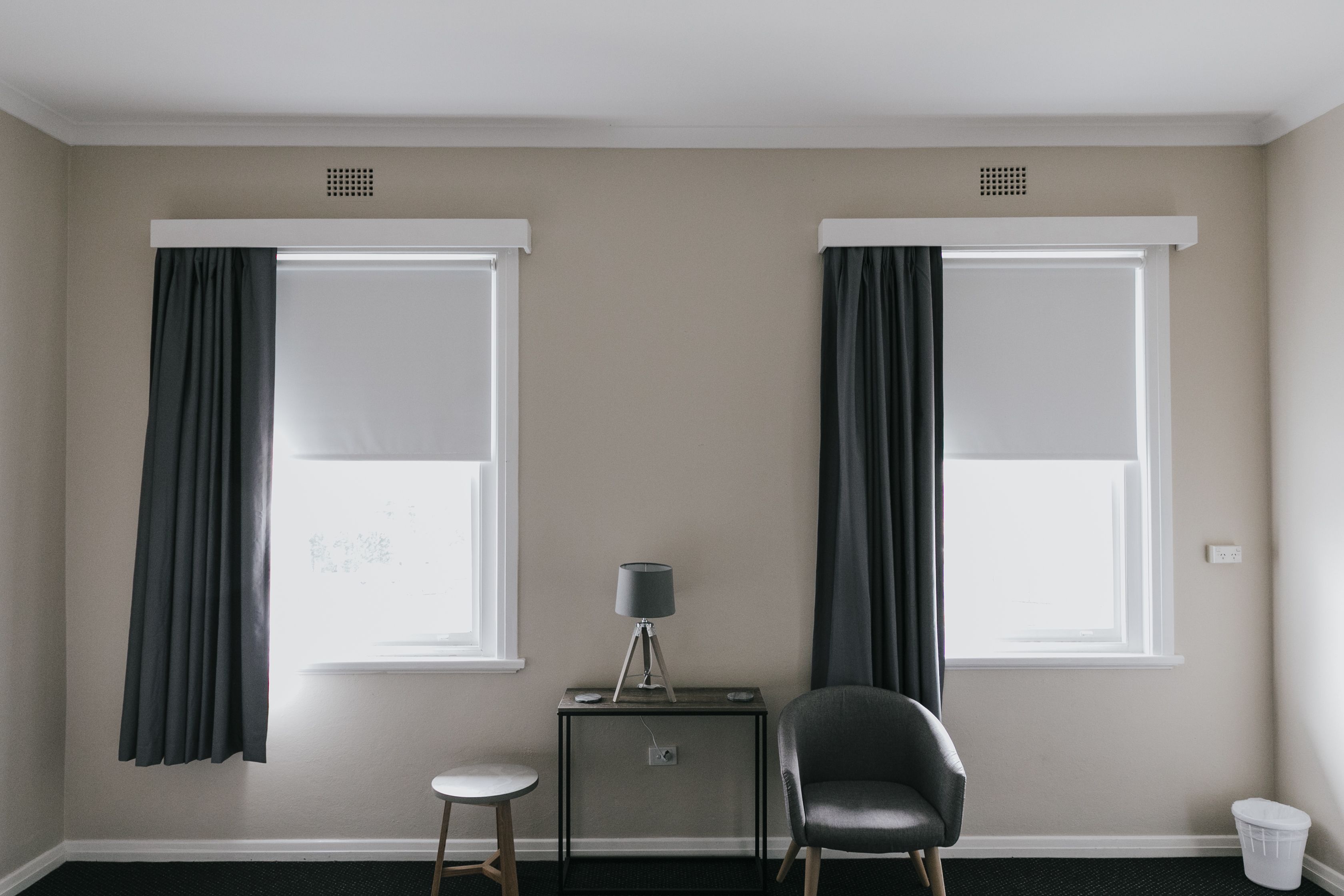 A room with two windows, a small table with a lamp, a chair, and a trash bin.