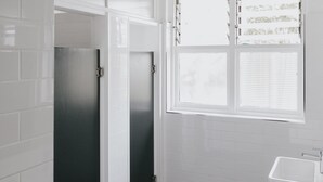 Une salle de bain avec des murs recouverts de carreaux blancs, un sol carrelé de noir, une fenêtre avec des stores et un lavabo blanc.