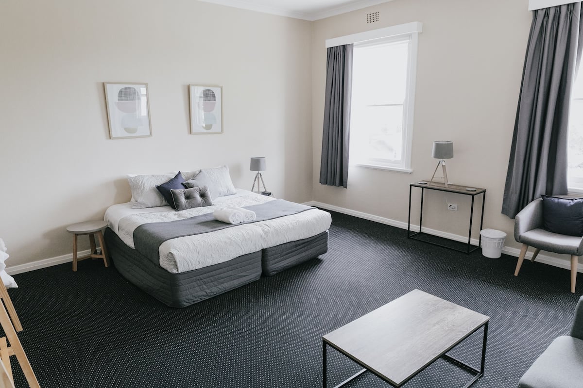 Hotellihuone, jossa on suuri sänky, pieni pöytä, tuoli ja ikkuna, jossa on verhot.