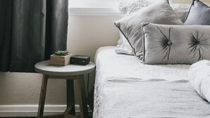 Une chambre à coucher avec un lit, des oreillers, une petite table sur laquelle se trouve une plante, et une fenêtre avec des rideaux.