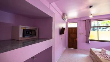 Una habitación con pared rosa, una cama con manta, un microondas, una puerta de madera y una ventana con cortinas.