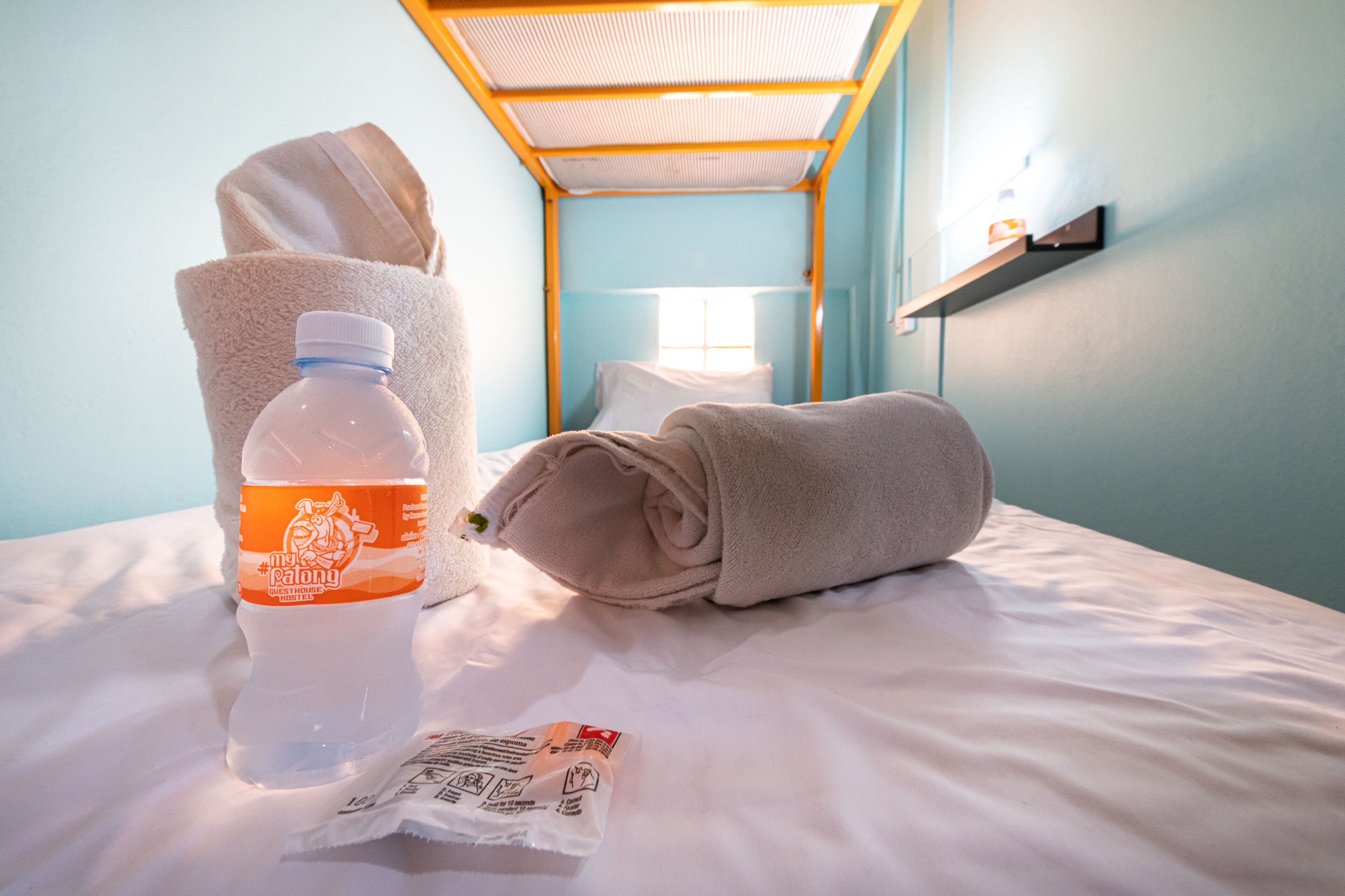 Una cama bien tendida con una toalla enrollada, una botella de agua y pañuelos desechables.
