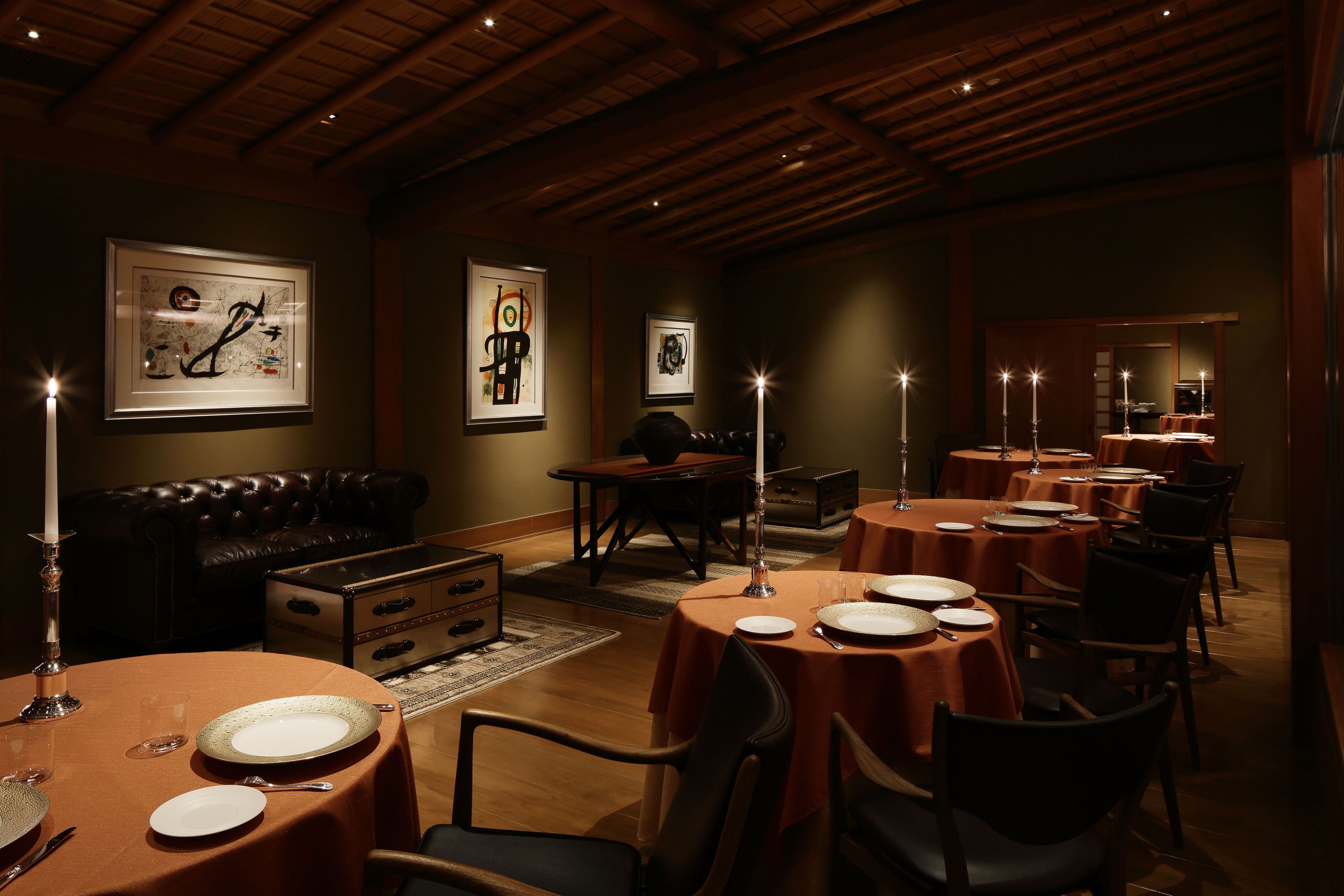 A dimly lit dining area with round tables, candles, and framed artwork on the walls.