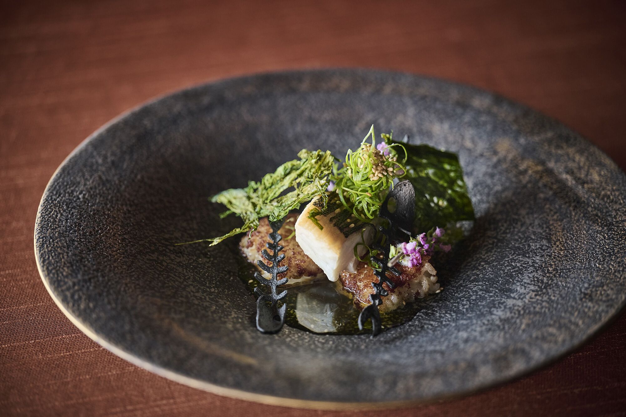 Un plato de cerámica oscura con un guiso de pescado, guarnición verde y un adorno sobre una superficie texturizada.