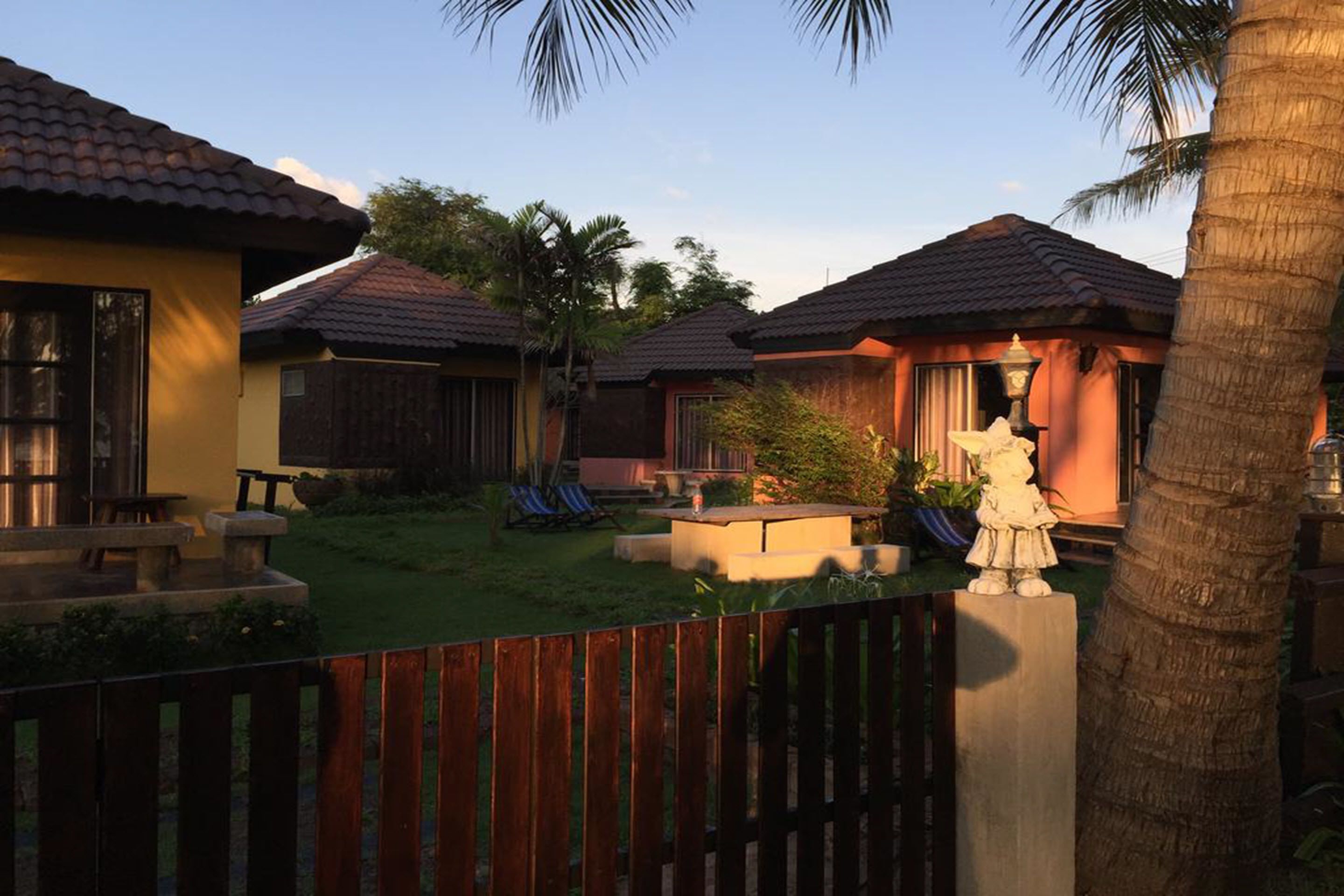 A resort with multiple villas, each with a tiled roof, a small patio, and a lawn. There are palm trees and a statue in the foreground.