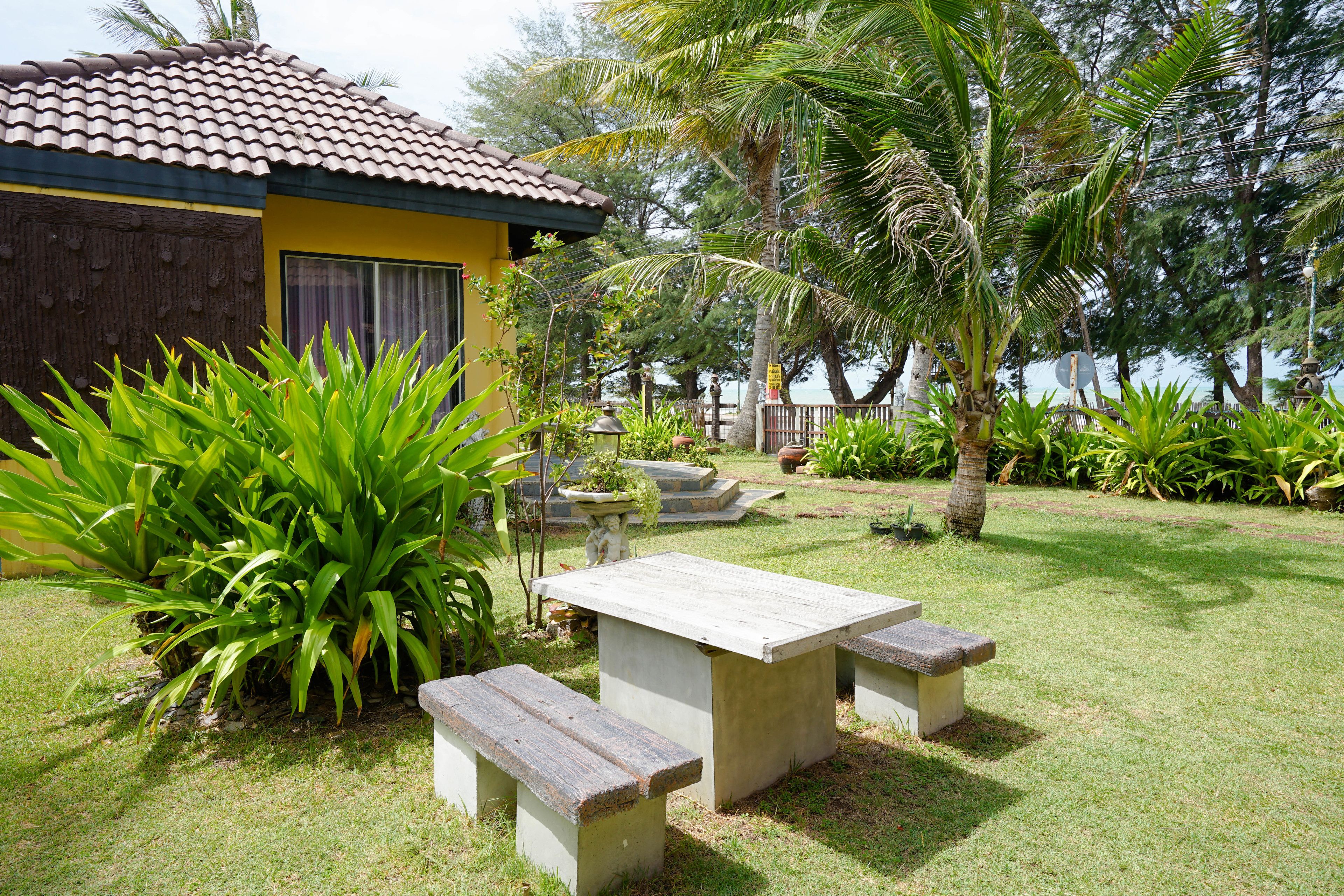 A patio with a table and benches, surrounded by lush greenery and a yellow building with a tiled roof.