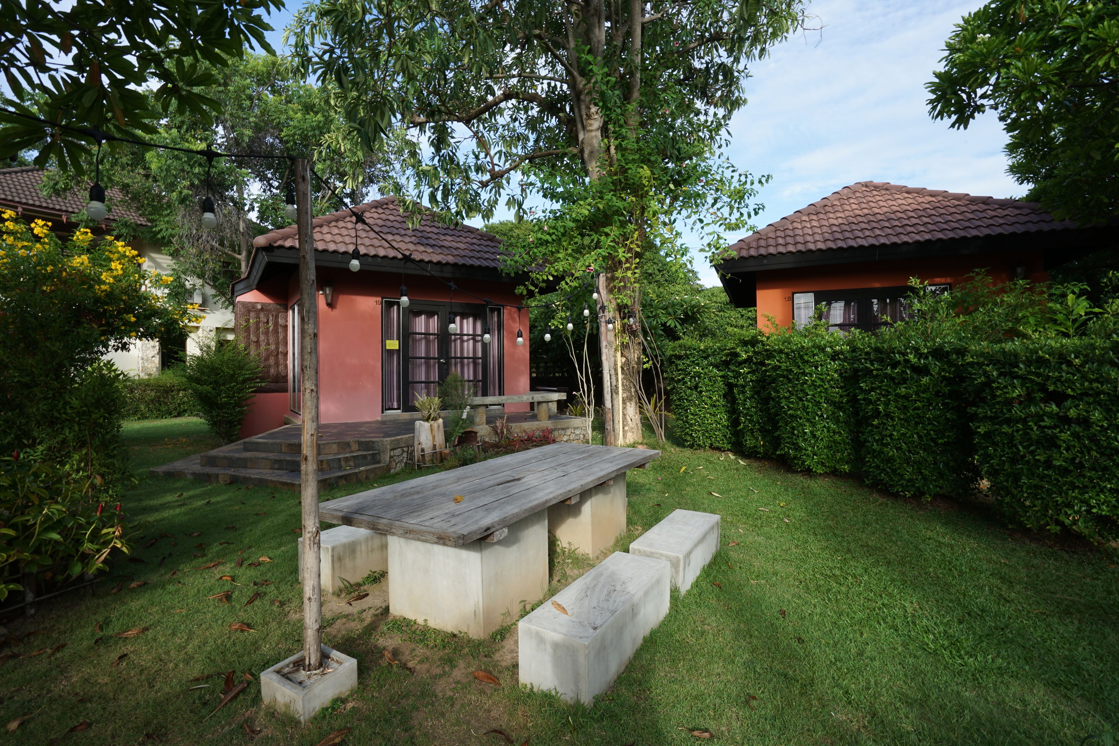 A courtyard with a wooden table and benches, surrounded by greenery and two buildings with red-tiled roofs.