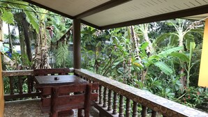 Uma área externa coberta com uma mesa e cadeiras de madeira, rodeada por vegetação exuberante e bananeiras.