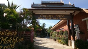 A entrada do Resort Baan Meak Lom, com uma placa em tailandês, rodeada por vegetação exuberante e palmeiras.