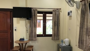 Quarto pequeno com mesa de madeira, dois banquinhos, geladeira, ventilador e janela com cortinas.