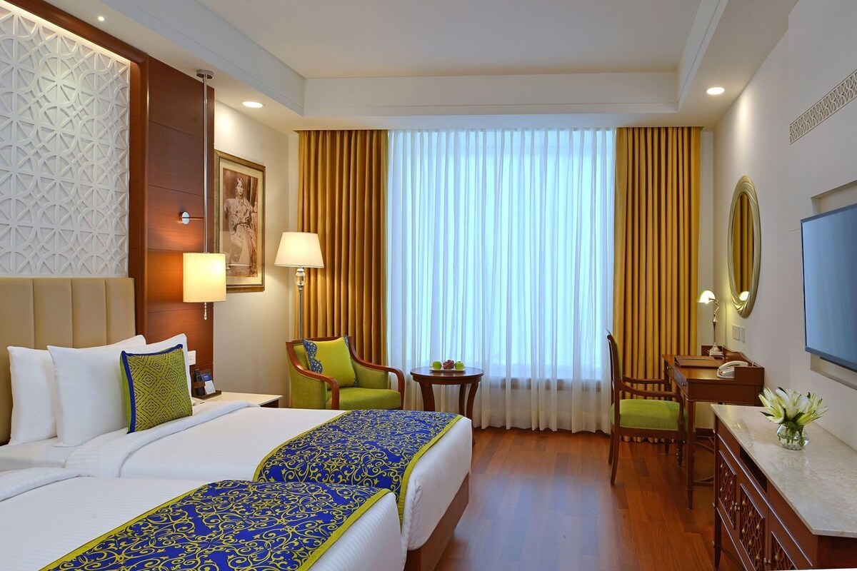 A hotel room with two beds, a desk, a chair, a TV, and a large window with curtains.