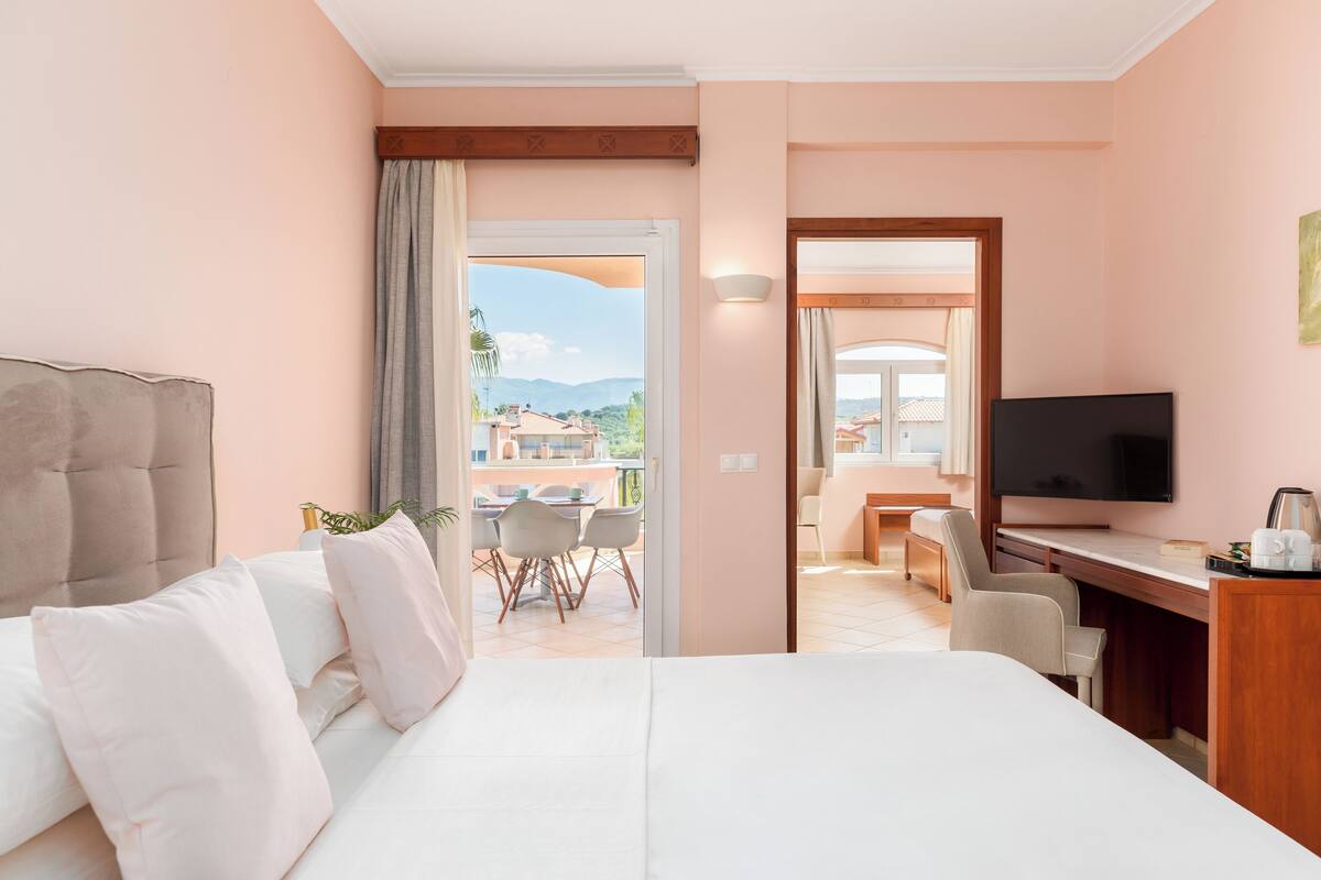 A hotel room with a bed, a desk, a TV, and a balcony with a view of the city.