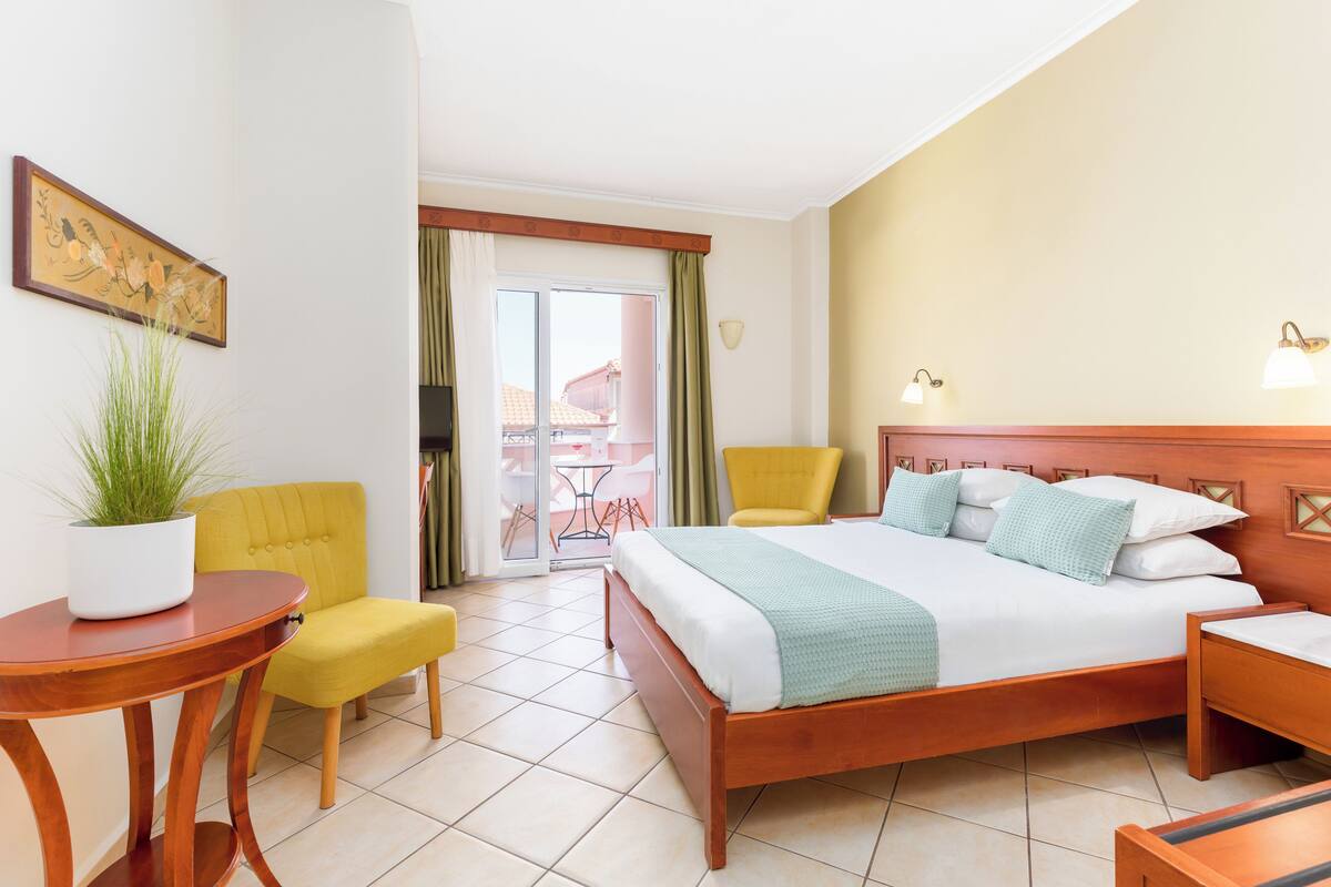 A hotel room with a bed, a yellow armchair, a wooden side table, and a potted plant.