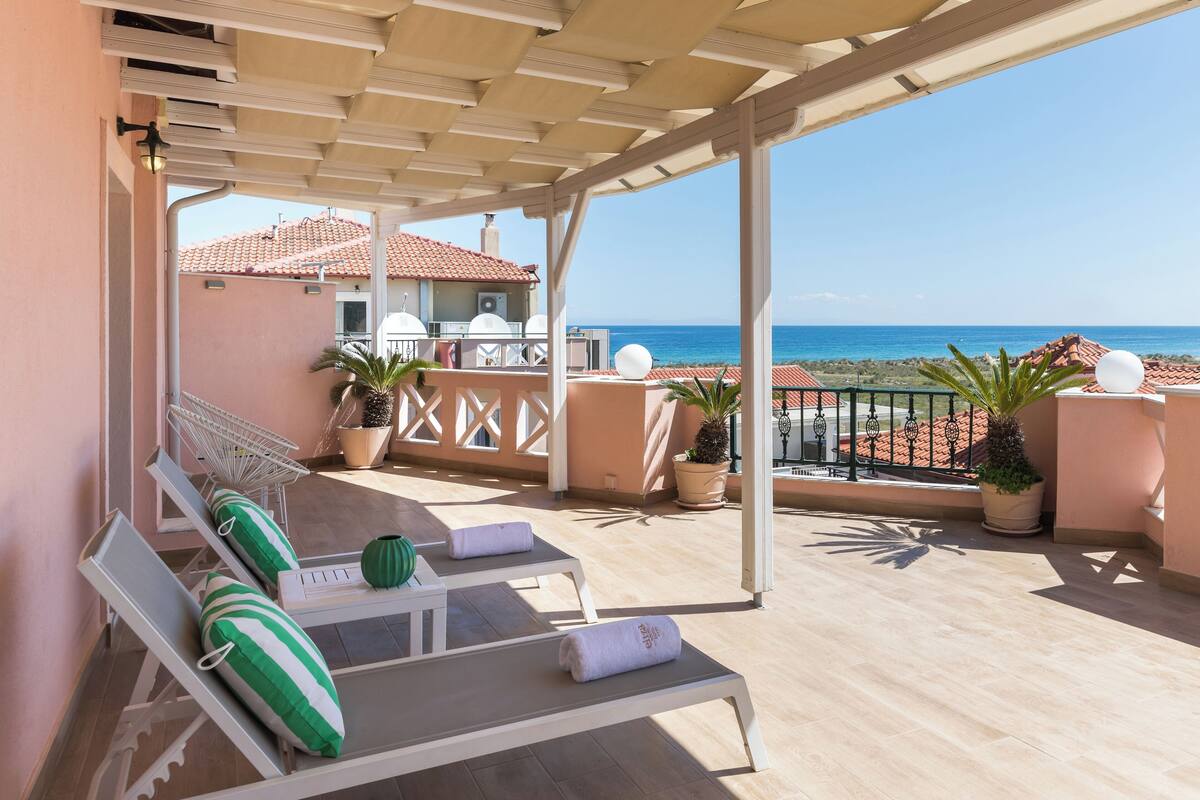 A covered outdoor seating area with lounge chairs and potted plants, overlooking a coastal view.